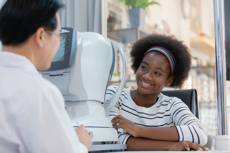 Niña sonriendo junto a un equipo de examen ocular con una profesional de la visión